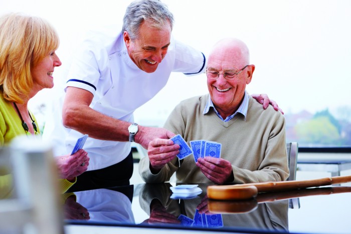 Senior man and woman play cards outdoors while a nurse watches. Horizontal shot.